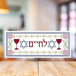 May include: A rectangular cross-stitch design with the Hebrew word "L'Chaim" in red, flanked by two Star of David symbols and wine glasses. The border features floral and geometric patterns in blue, green, and pink.