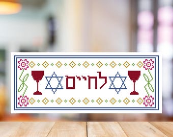 May include: A rectangular cross-stitch design with the Hebrew word "L'Chaim" in red, flanked by two Star of David symbols and wine glasses. The border features floral and geometric patterns in blue, green, and pink.