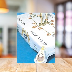 May include: White tablecloth with embroidered floral and Hebrew text. Features a Star of David and a wine bottle design. Two brass candlesticks are on the table. The tablecloth is on a blue surface.
