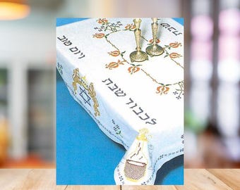 May include: White tablecloth with embroidered floral and Hebrew text. Features a Star of David and a wine bottle design. Two brass candlesticks are on the table. The tablecloth is on a blue surface.