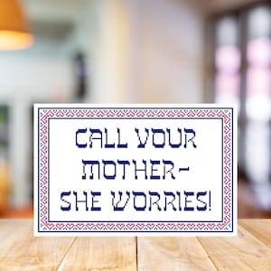 May include: A rectangular cross-stitch design with the text "CALL YOUR MOTHER - SHE WORRIES!" in navy blue. The text is framed by a border of navy blue and red cross-stitch patterns. The sign is on a wooden surface.