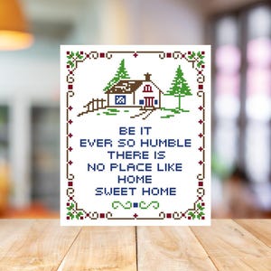 May include: A cross-stitch style artwork featuring a house and trees, with the text "BE IT EVER SO HUMBLE THERE IS NO PLACE LIKE HOME SWEET HOME" in blue. The border has green and red floral accents. The artwork is displayed on a wooden surface.