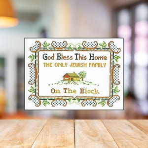 May include: A framed cross-stitch with the text "God Bless This Home, The Only Jewish Family On The Block." The design features a house illustration and decorative border with green and brown accents. The background is blurred.