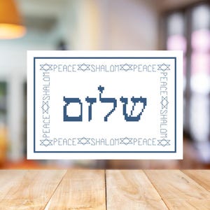 May include: A framed cross-stitch design with the Hebrew word "Shalom" in blue. The design is surrounded by a border with the words "Peace" and "Shalom" and Star of David symbols. The background is blurred.