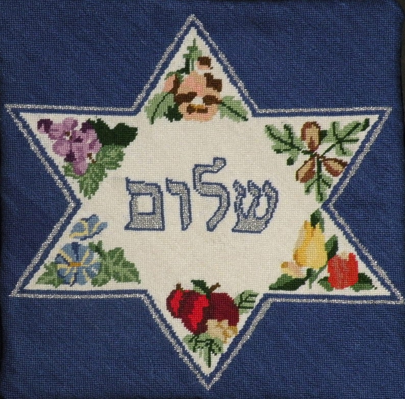 K&ouml;nnte beinhalten: Ein wei&szlig;er gestickter Davidstern mit silbernem Rand. Der Stern ist mit bunten Blumen und Fr&uuml;chten verziert. Das hebr&auml;ische Wort "Shalom" ist in Silber in der Mitte des Sterns gestickt.