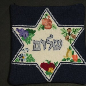 K&ouml;nnte beinhalten: Ein wei&szlig;er Stoff-Davidstern mit silbernem Umriss, bestickt mit dem hebr&auml;ischen Wort &bdquo;Shalom&ldquo; und verschiedenen Fr&uuml;chten und Blumen. Der Stern ist von einem dunkelblauen Stoff umgeben.