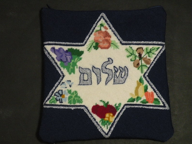 K&ouml;nnte beinhalten: Ein wei&szlig;er Stoff-Davidstern mit silbernem Umriss, bestickt mit dem hebr&auml;ischen Wort &bdquo;Shalom&ldquo; und verschiedenen Fr&uuml;chten und Blumen. Der Stern ist von einem dunkelblauen Stoff umgeben.