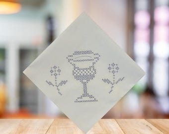 May include: White square cloth with a cross-stitch design featuring a chalice and two floral motifs in dark blue. The cloth is displayed on a wooden surface, with a blurred background.