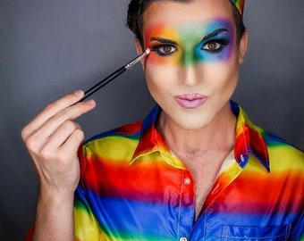 May include: A person wearing a rainbow striped shirt and a rainbow crown with colorful makeup on their face. They are holding a makeup brush near their eye.