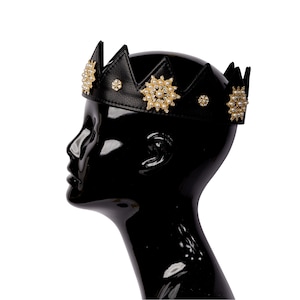 Puede incluir: Una corona de cuero negro con detalles dorados y adornos de perlas. La corona se lleva en una cabeza de maniquí negra.