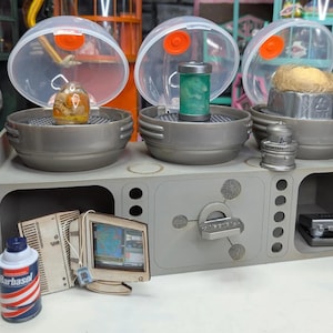 Könnte beinhalten: Ein graues, miniaturisiertes Labor mit drei transparenten Kuppeln, die jeweils einen anderen Gegenstand enthalten. Eine Dose Barbasol-Rasiercreme und ein Miniaturcomputer befinden sich im Vordergrund. Das Setup hat eine retro-futuristische Ästhetik.