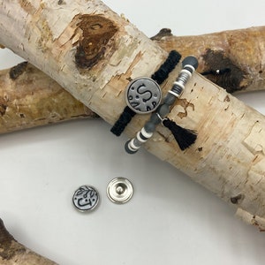 May include: Two bracelets with black and white beads and a silver charm with the letter "S" on a birch log background. Two additional silver charms are visible in the foreground.