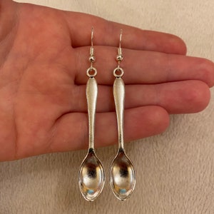 May include: A pair of silver spoon earrings. Each earring has a miniature spoon design with a hook for pierced ears. The spoons have a reflective, polished finish, and the earrings are displayed against a neutral background.