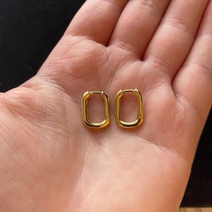 May include: A pair of gold-coloured, rectangular hoop earrings. The earrings have a smooth, polished finish and a simple, modern design. The earrings are shown on a light-coloured background.