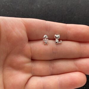 May include: A pair of silver-colored stud earrings. One earring is shaped like a fish skeleton, and the other is a small cat. The earrings are small and delicate, perfect for everyday wear.
