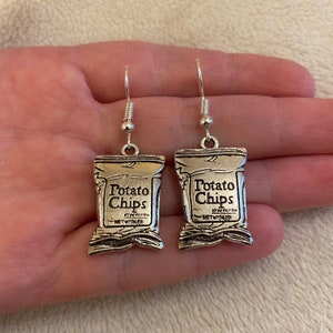 Może przedstawiać: Srebrne kolczyki wiszące w kształcie torebki chipsów ziemniaczanych z napisem "Potato Chips" z przodu.