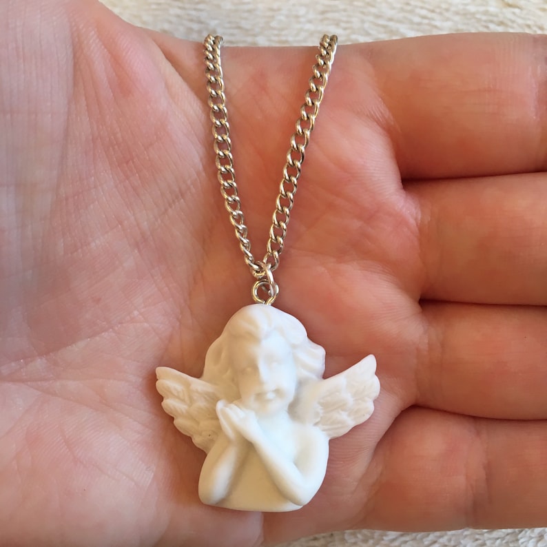Op de afbeelding: Een witte porseleinen engelhanger met vleugels, handen gevouwen in gebed, hangend aan een zilveren ketting.