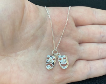 May include: A silver necklace with a pendant featuring two silver masks, one with a smiling face and the other with a frowning face.