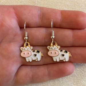 Op de afbeelding: Een paar zilveren oorhangers met gouden koe-bedels. De koeien zijn wit met zwarte vlekken en roze neuzen.