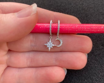 May include: A pair of silver hoop earrings with a dangling star and crescent moon charm. Both charms are encrusted with small, clear cubic zirconia stones.