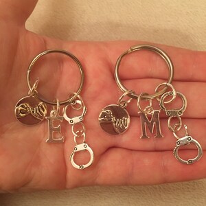 May include: Two silver keychains with a silver charm of two hands clasped together, a silver letter charm, and two silver handcuff charms. The keychains are labeled with the letters "E" and "M".