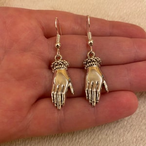 Op de afbeelding: Een paar zilveren oorhangers met een gedetailleerd handontwerp. De handen zijn open met gestrekte vingers en hebben een getextureerde manchet aan de pols.