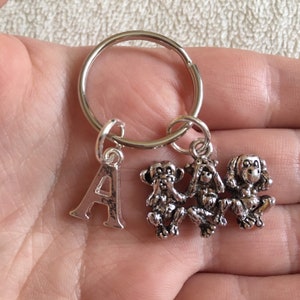 May include: Silver keyring featuring three monkeys, one covering its eyes, one covering its ears, and one covering its mouth. The monkeys are attached to a silver ring with a silver letter 'A' charm.