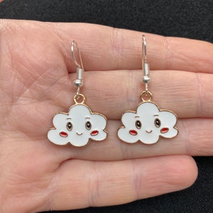 May include: A pair of cloud-shaped earrings. Each earring features a white cloud design with a gold outline, black eyes, and red lips. The earrings have silver-coloured fish hook ear wires.