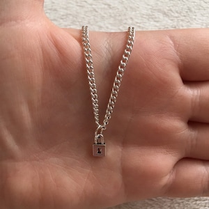 Op de afbeelding: Zilverkleurige ketting met een kleine hangslot-hanger. De ketting is een gourmette-ketting en de hanger heeft een sleutelgatontwerp. De ketting wordt getoond tegen een neutrale achtergrond.
