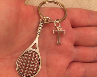 May include: Silver-toned tennis racket keychain with a brown net and a letter "T" charm. The keychain has a gold-colored ring for attaching keys. A sporty accessory for tennis enthusiasts.