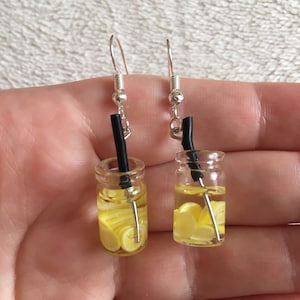 May include: A pair of dangle earrings featuring miniature glass jars filled with yellow lemon slices and a black straw. The jars are suspended from silver hooks.