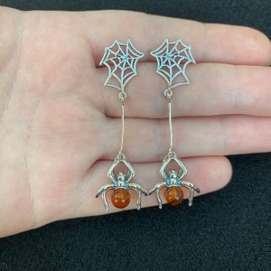 Op de afbeelding: Een paar zilveren oorhangers met een spin met een amberkleurige edelsteen en een webontwerp.
