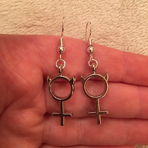 Könnte beinhalten: Ein Paar silberne Ohrringe mit einem weiblichen Symbol-Design. Jeder Ohrring hat einen kleinen Kreis mit Hörnern oben und einem Kreuz unten.