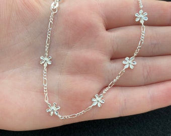 May include: A silver chain necklace with five small flower charms. The necklace is on a hand.