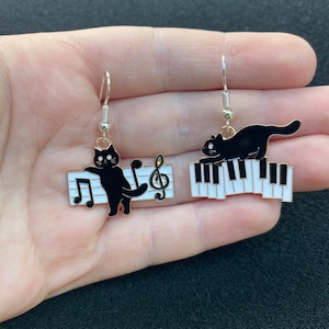 Puede incluir: Un par de pendientes de gato negro con un diseño de teclado de piano. Un gato está tocando una nota musical en el piano, mientras que el otro gato está sentado en el piano.
