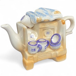 Puede incluir: Una caprichosa tetera de cerámica con forma de casa, pintada en crema, beige y azul. La tetera tiene un pico, un asa y una tapa con un diseño de rayas azules y blancas. También se incluyen dos tazas pequeñas y dos cuencos.