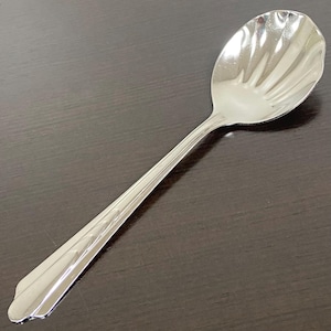 Può includere: Un cucchiaio in metallo color argento lucido con una coppa dal design a conchiglia smerlata e un manico affusolato con motivo a coste. La superficie riflettente del cucchiaio mostra riflessi di luce lungo i bordi della coppa e del manico.