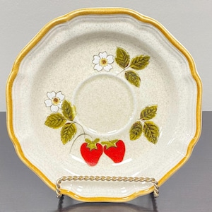Può includere: Un piattino in ceramica rotondo con bordo smerlato e finitura color crema maculato. Il bordo è accentuato da una fascia marrone dorato. Il centro presenta un'illustrazione botanica di due fragole rosse, foglie verdi e fiori bianchi.