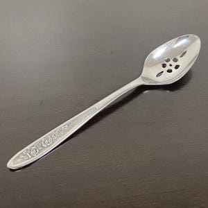 Può includere: Un cucchiaio da portata forato in metallo color argento con finitura lucida. La parte concava presenta un motivo decorativo a forma di fiore, mentre il manico è decorato con un disegno floreale in rilievo su uno sfondo testurizzato.