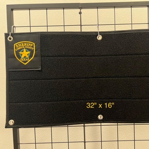 Puede incluir: Organizador táctico negro con el texto "32" x 16"" y un parche que dice "SHERIFF LUBBOCK COUNTY TEXAS". El organizador tiene múltiples tiras de velcro horizontales y ojales metálicos. Mide 81 cm x 41 cm.