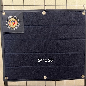 Puede incluir: Un panel de pared rectangular de tela azul oscuro con una superficie de velcro. Tiene un parche con el logotipo de Lubbock Fire Rescue y el texto "24" x "20".