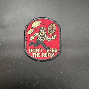 May include: Embroidered patch with a red background and a black border. The patch features a cartoon referee running with coins and a smiling face. Text on the patch reads "DON'T FEED THE REFS!"