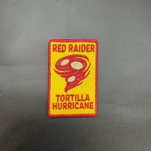 May include: A rectangular patch with a yellow background and red border. The patch features the words "RED RAIDER" at the top and "TORTILLA HURRICANE" at the bottom in red lettering. A red and white tornado graphic is in the centre.