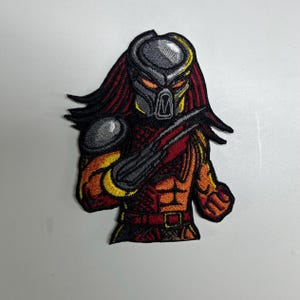 May include: Embroidered patch depicting a Predator character. The patch features the Predator's helmet, dreadlocks, and armour in grey, red, orange, and yellow. The design is detailed with intricate stitching, creating a textured appearance.