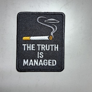 May include: Black rectangular patch with white embroidered text that reads "THE TRUTH IS MANAGED". The patch features a lit cigarette with smoke rising and a UFO above it.