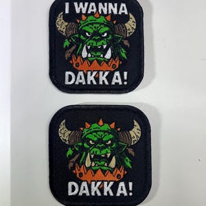 May include: Two black embroidered patches with a green monster face, brown horns, and the text "I WANNA DAKKA!". The monster has orange accents and white teeth. The patches are square with rounded corners.
