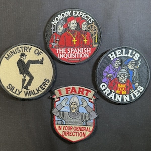 May include: Four circular and shield-shaped embroidered patches featuring colorful illustrations and text. Designs include a figure walking with a cane, three figures in red robes, three elderly figures, and a knight with the text I FART IN YOUR GENERAL DIRECTION.