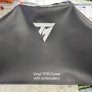 May include: A dark gray vinyl cover with a white logo and the text "Vinyl TPR Cover with embroidery". The cover is draped over an unseen object, with a textured surface and a stitched edge.