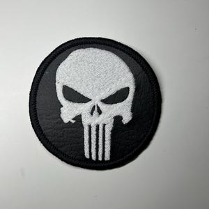 Könnte beinhalten: Ein runder, schwarzer Aufnäher mit einem weißen Totenkopf-Emblem. Der Totenkopf hat Augenhöhlen und eine Nasenhöhle, mit vertikalen Linien darunter. Der Aufnäher ist wahrscheinlich aus Stoff mit einem genähten Rand.