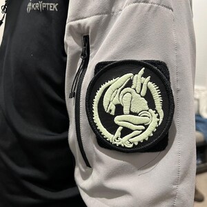 Op de afbeelding: Een zwart-grijze jas met een Kryptrek-logo en een ronde patch met een gloeiend groen alien-ontwerp. De jas heeft een zwart zakje met ritssluiting op de mouw.